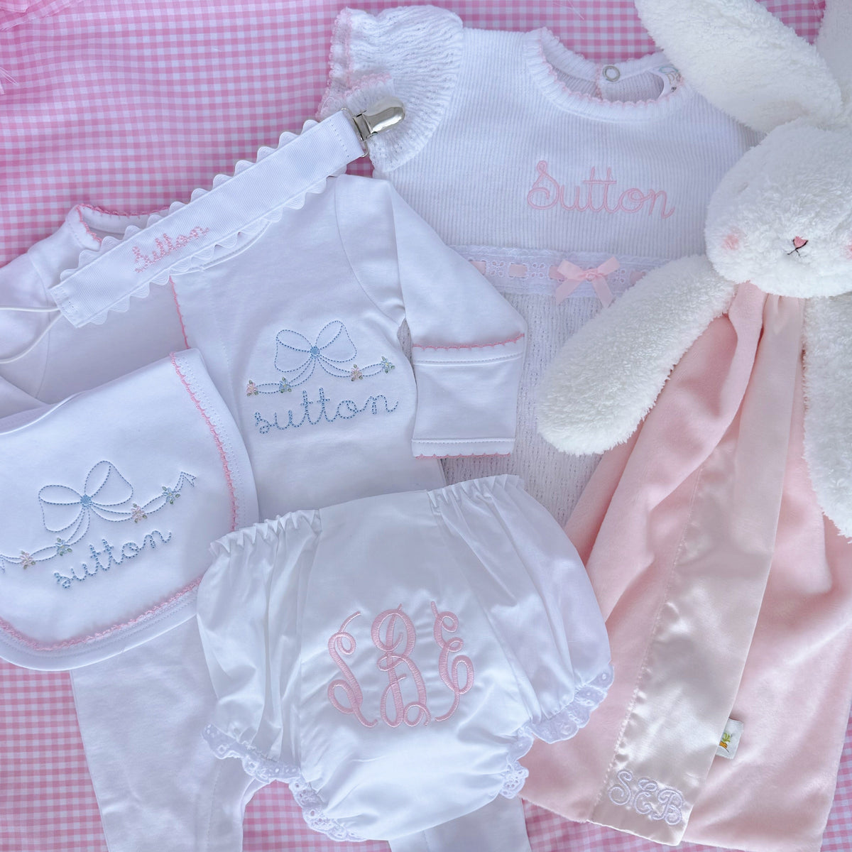 A baby gift set arranged on pink gingham fabric, including a white long-sleeve romper with pink embroidery, matching white diaper cover, white bib, white diaper, and a plush white bunny toy with a pink blanket. The word 'sutton' is embroidered on several items.